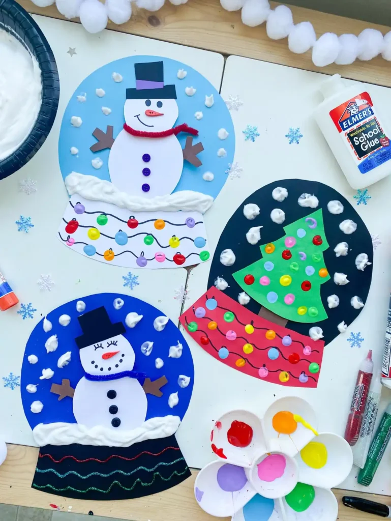 Three paper snow globes with a snowman, a green tree and fingerprint light string bases