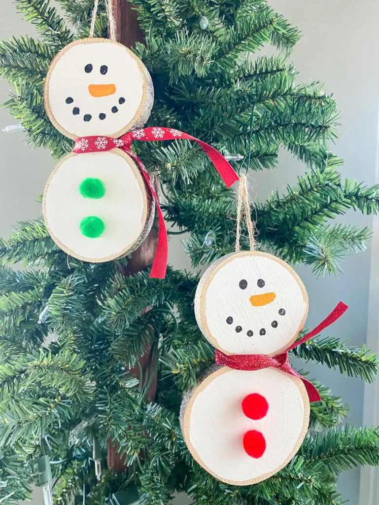 Two wood slice snowman ornaments with ribbon scarves and pom pom buttons hanging on a christmas tree