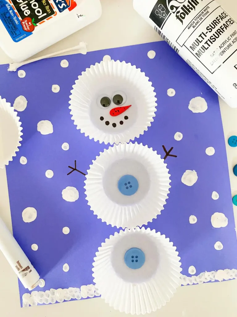 Cupcake liner snowman glued on purple paper with googly eyes, an orange nose and blue buttons