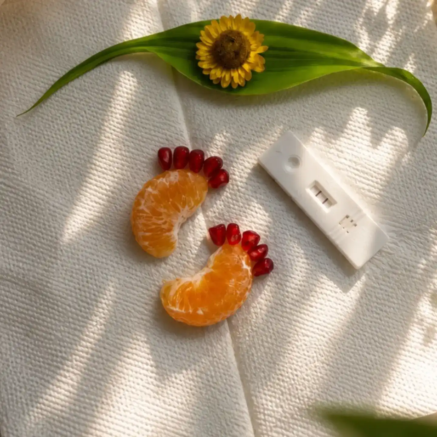 Pregnancy announcement to husband idea: baby feet made from two mandarin segments and pomegranate seeds beside a pregnancy test on a cream woven cloth with a small sunflower on a green leaf