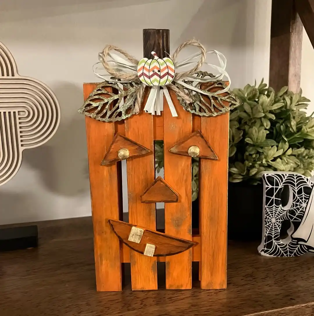 Pumpkin made of bold orange slats with a dark stem, a triangle face with gold tack eyes and a crescent smile with two tan square teeth, topped with twine, ribbon and leaf shapes like a bow