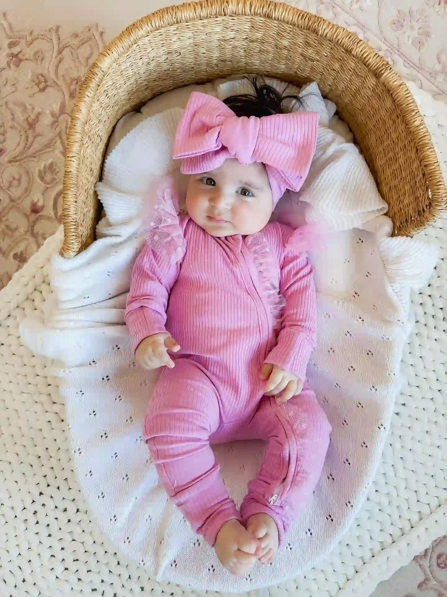 Baby sleep gowns alternative: baby in a pink ribbed zip sleeper with a bow headband lying in a woven basket