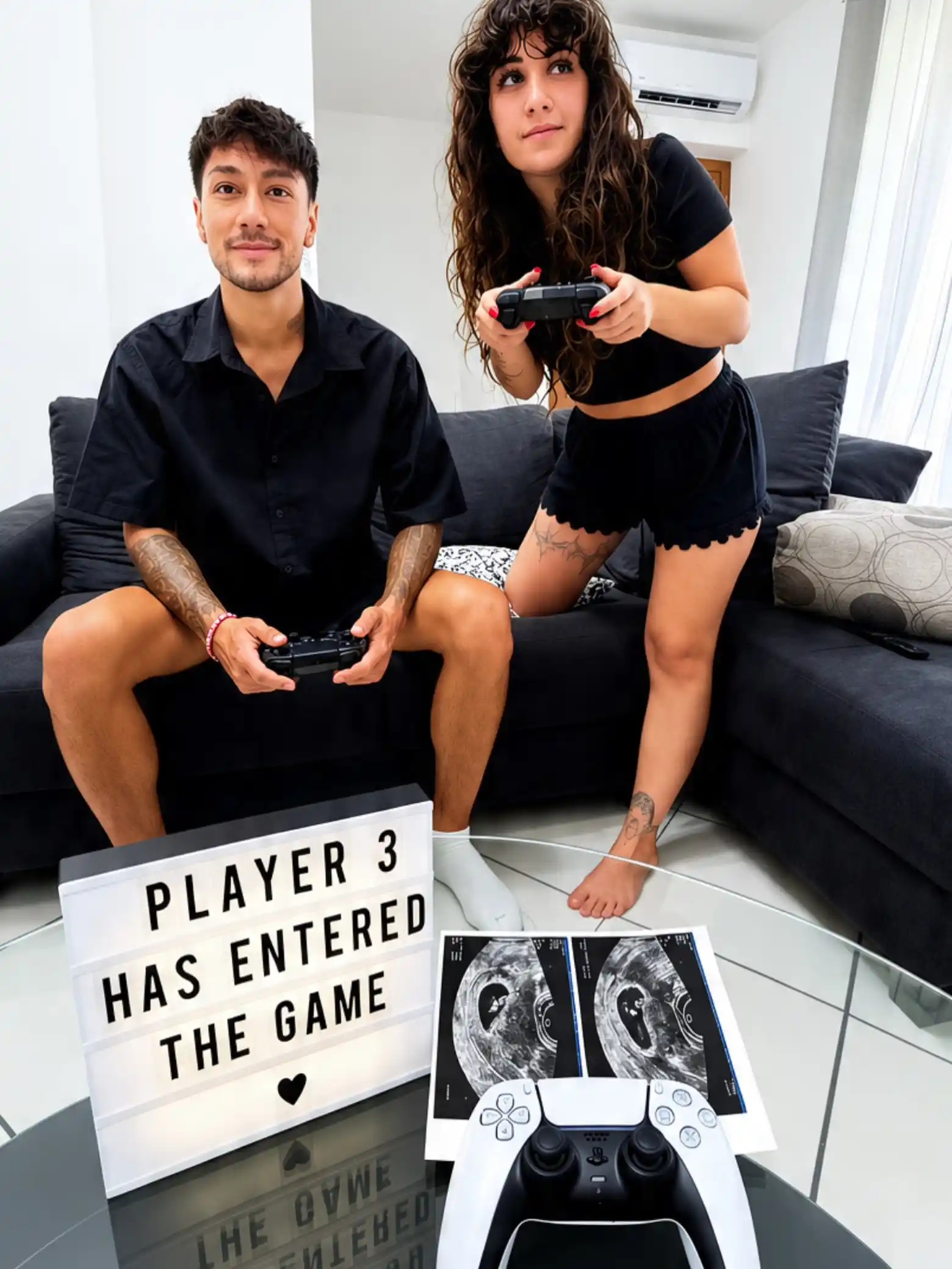 Couple in black outfits holding game controllers on a dark sofa with a light box reading "Player 3 has entered the game" and ultrasound photos on a glass table