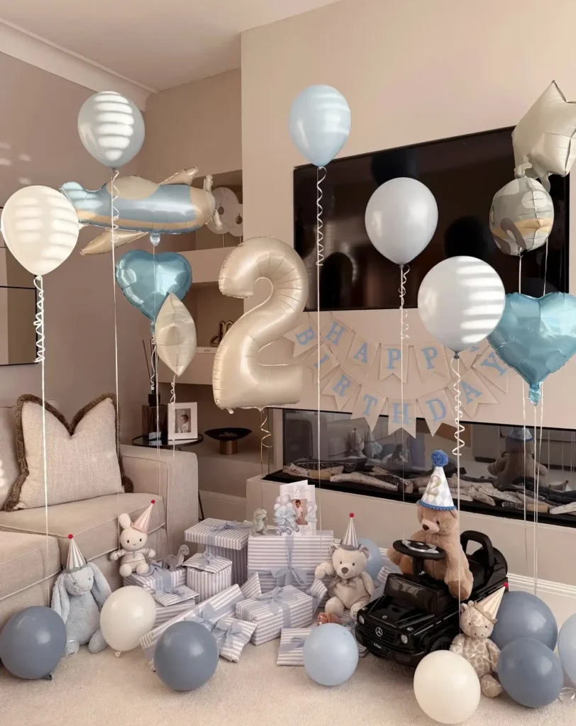 2nd birthday party ideas: living room birthday setup with pale blue and cream balloons, a plane foil, heart foils and a big cream number 2, a cream banner above the fireplace, striped gifts, plush toys in party hats and a black ride-on car with a teddy bear driver