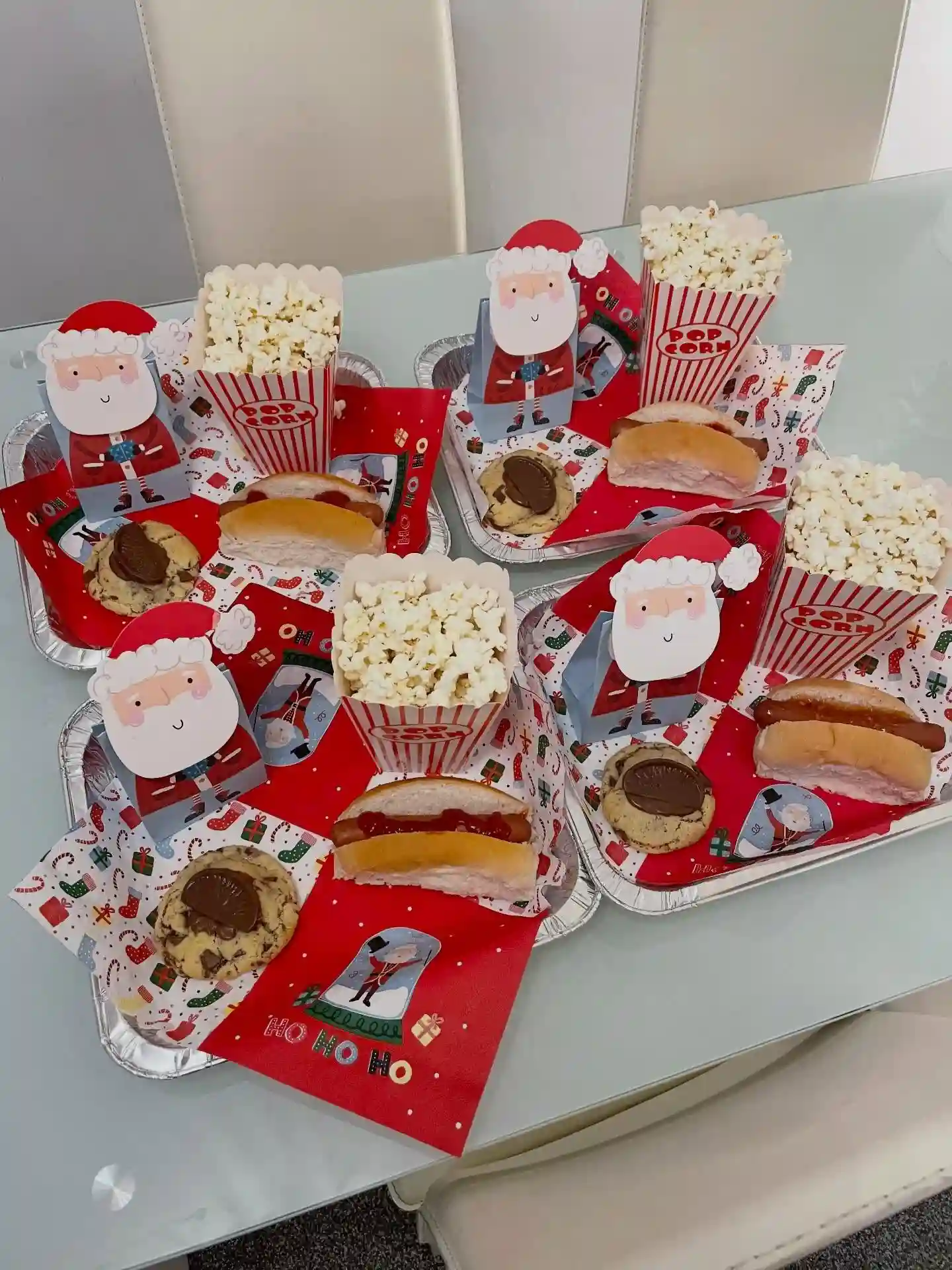 Four foil trays set on a glass table, each with a Santa gift bag, a striped popcorn box, a hot dog in a bun, a chocolate chip cookie, and Christmas napkins.
