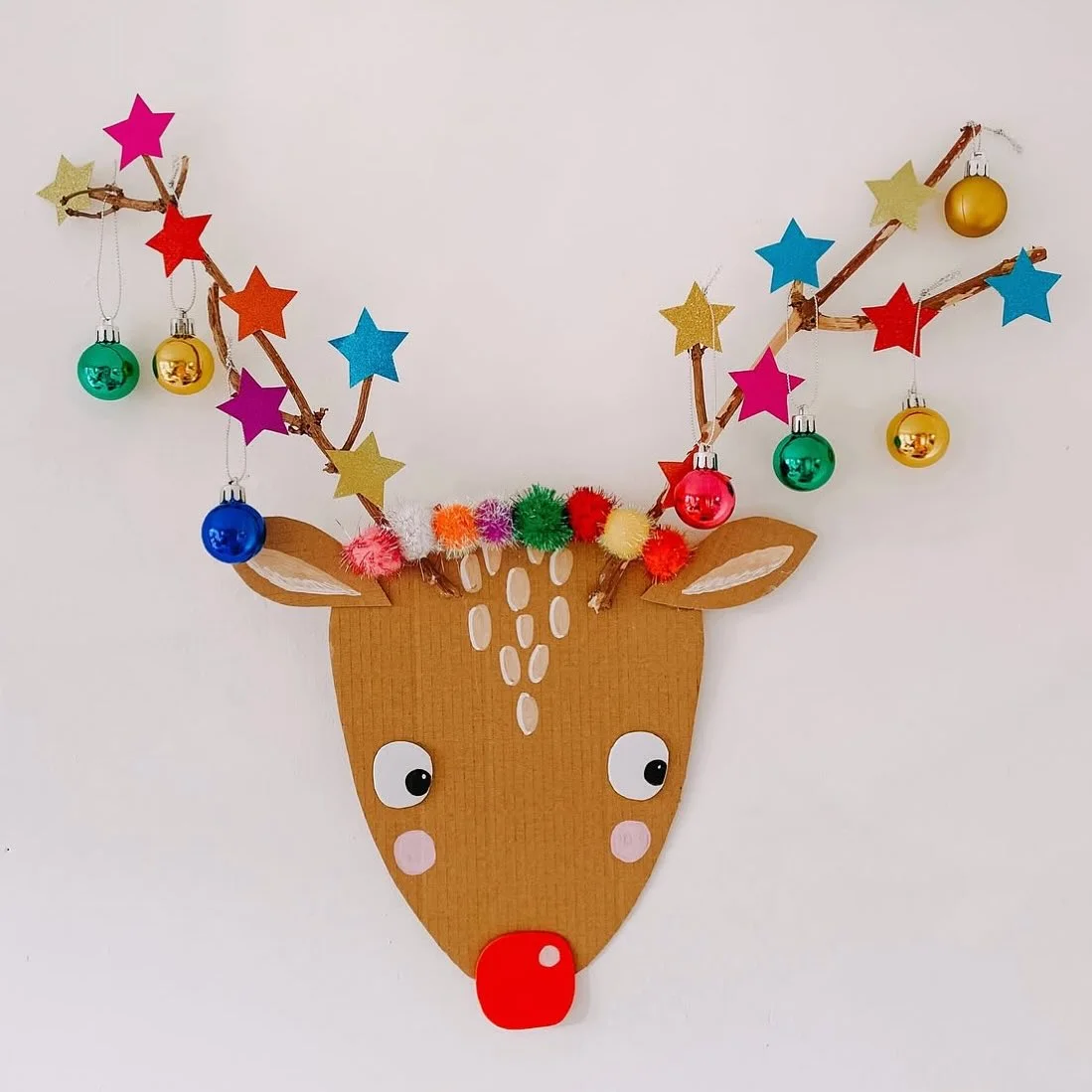 A brown cardboard reindeer face on a white wall with a red nose, pom pom fringe, and twig antlers covered in glitter stars and mini baubles.