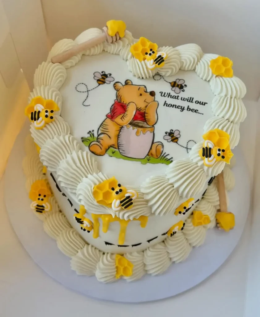 Heart shaped Winnie the Pooh gender reveal cake with piped cream border, yellow bees and honeycomb, and the words what will our honey bee