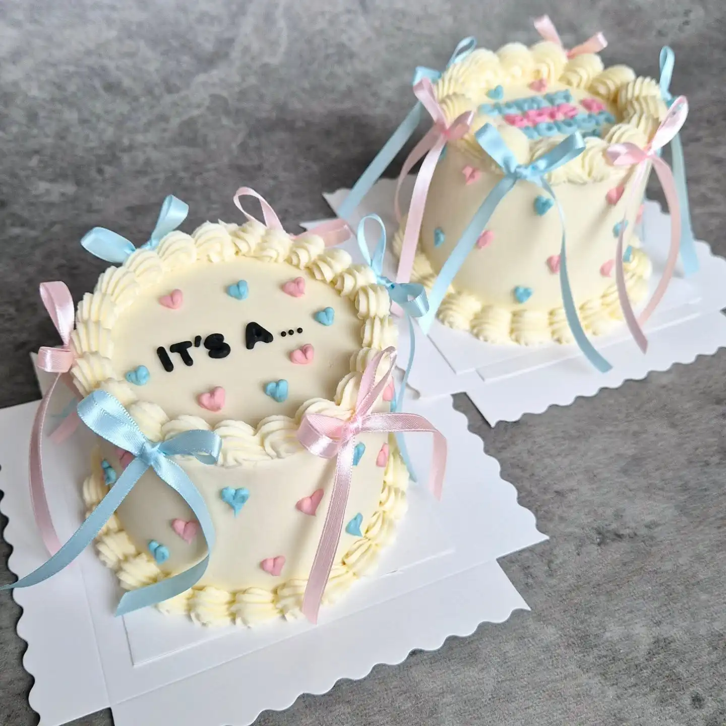 Two small cream gender reveal cakes with pink and blue piped hearts and satin ribbon bows, one with the words it's a in black letters