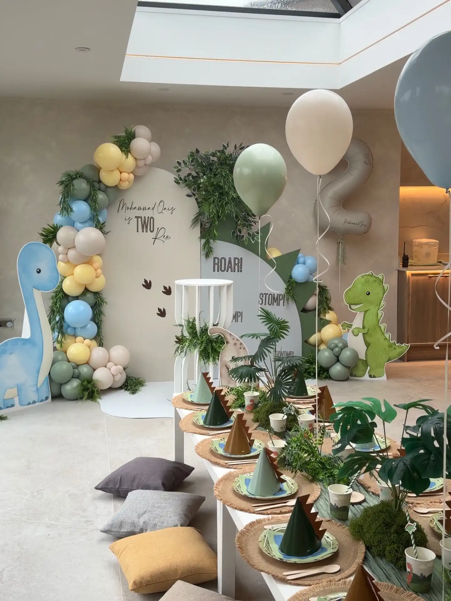 Dinosaur party backdrop with ROAR and STOMP wording and brown footprints, a blue long neck and a green T-rex cutout, a table with woven placemats, spiked paper party hats and moss, ferns and leaves, and low floor cushions