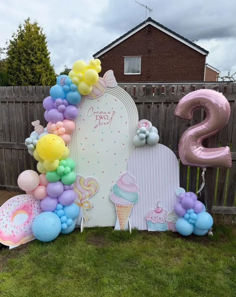 Candy corner birthday setup in front of a wooden fence with a white sprinkle dot arch, a pink striped arch, yellow, mint and lilac balloons, flat donut, lollipop, ice cream and cupcake cutouts and a big pink foil 2