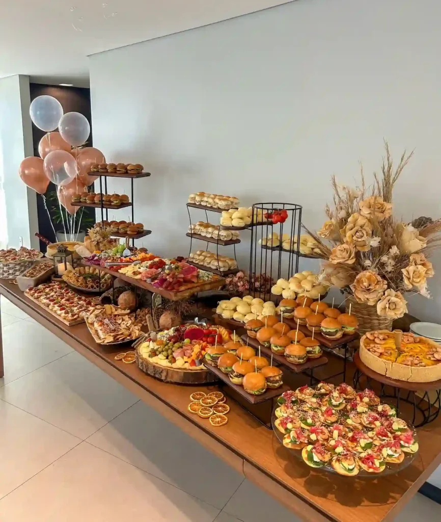 Long wooden party table with tiered stands of mini burgers and soft rolls, a charcuterie board, a savoury tart and rose gold balloons