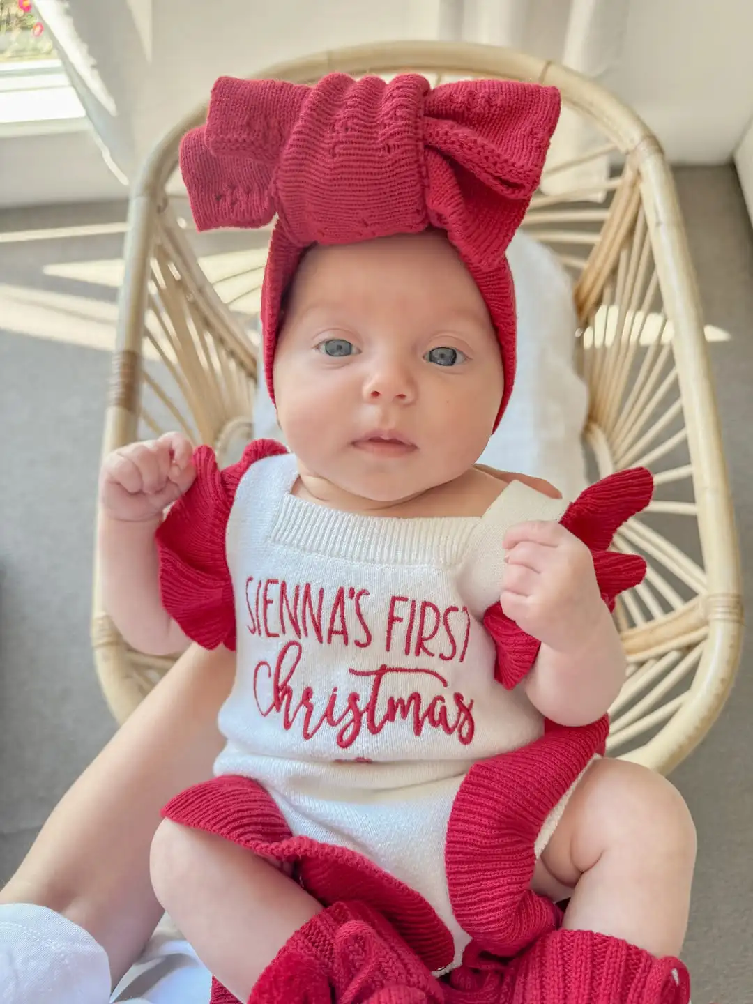 Baby first christmas ideas: baby in a cream knit romper with red ruffled sleeves and red script name lettering, red knit bow headband and leg warmers, sitting in a woven rattan basket by a bright window