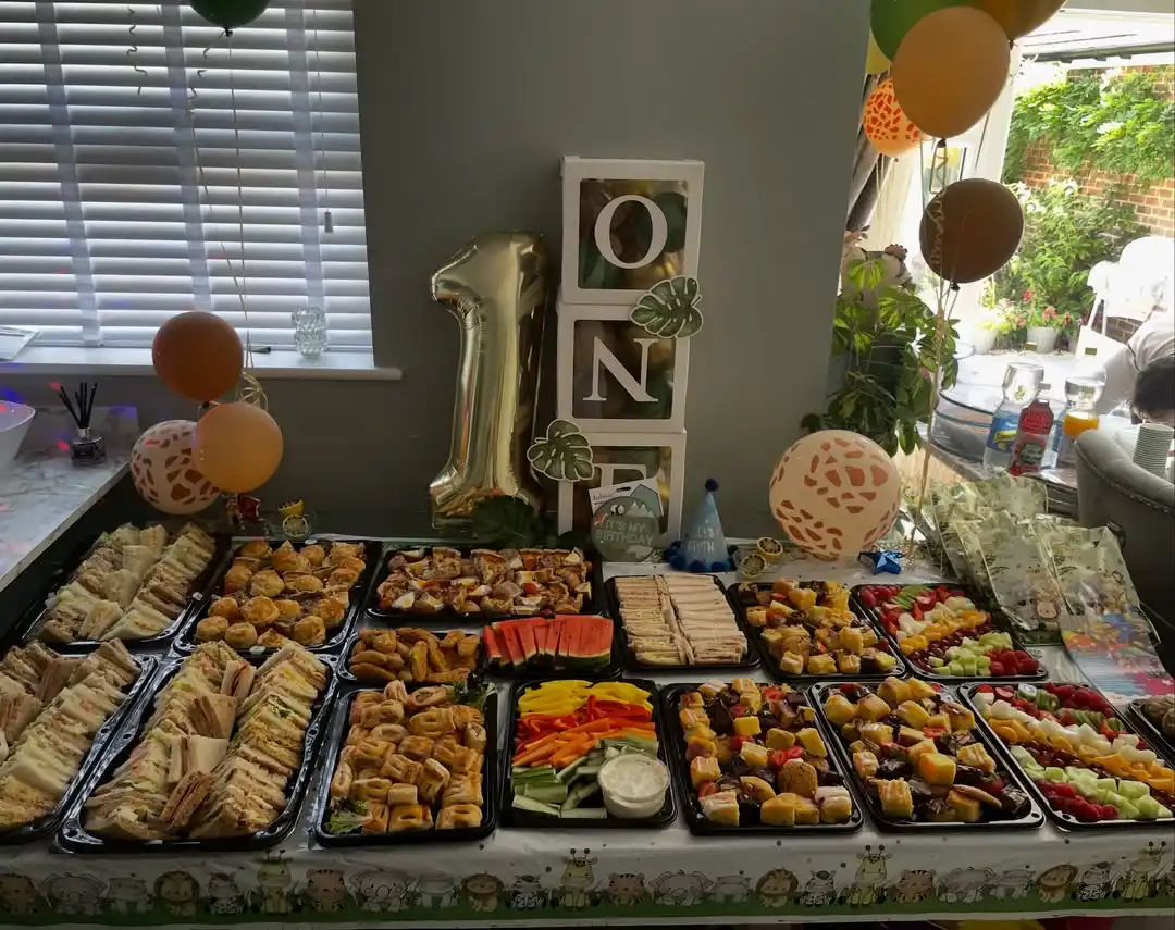 First birthday food table with black trays of sandwiches, pastry bites, fruit cubes, veggie sticks and dip under a safari tablecloth