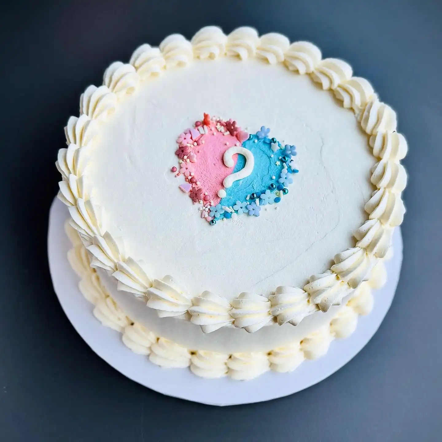 Round white buttercream cake with a shell border and a small pink and blue heart topped with a white question mark and sprinkles