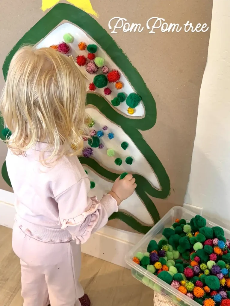 Toddler pressing pom poms onto a big christmas tree shape on the wall