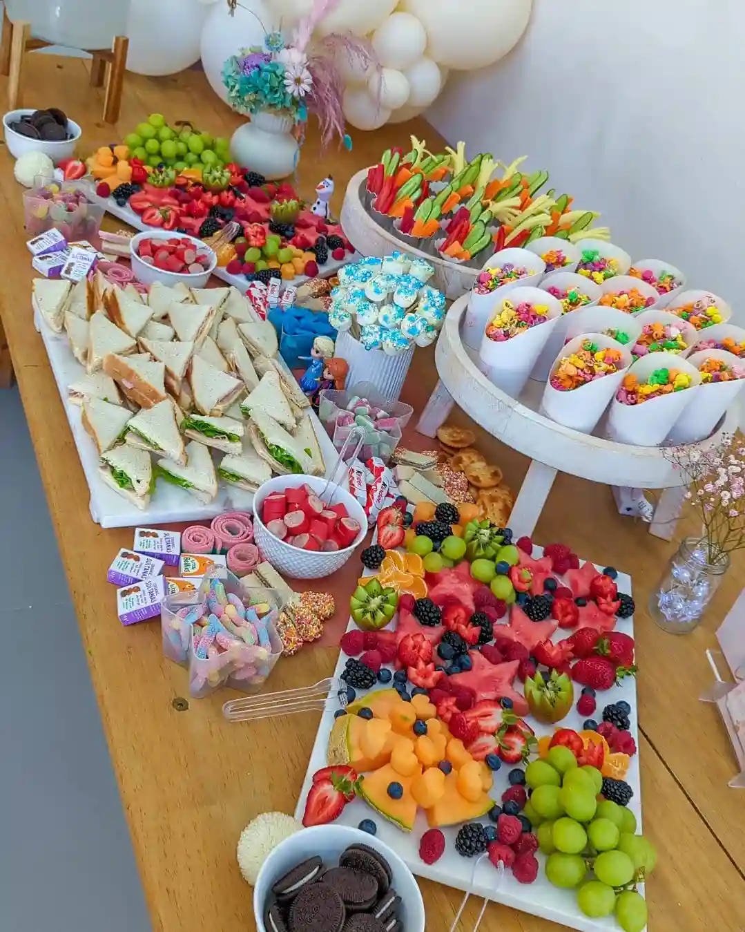 Icy blue and white birthday table with paper cones of colourful popcorn and veggie sticks, a fruit board and sandwich triangles