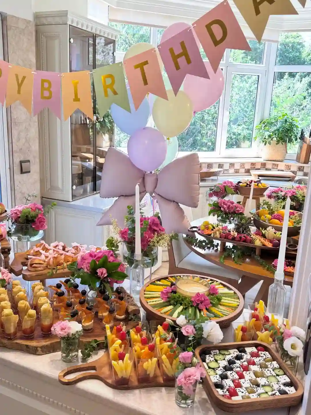 Pastel birthday table with finger food in clear cups, a veggie ring around a dip, a checkerboard tray and a bow balloon