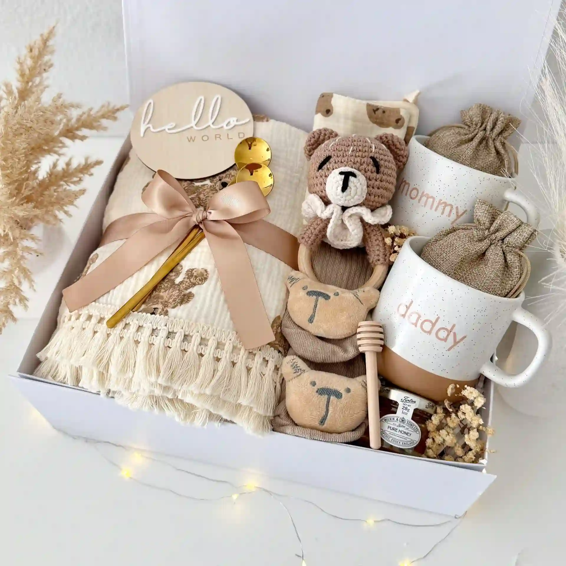 Gift box with speckled mommy and daddy mugs, a small jar of honey with a wooden dipper, a fringed muslin blanket, a crochet bear rattle and a wooden Hello World disc