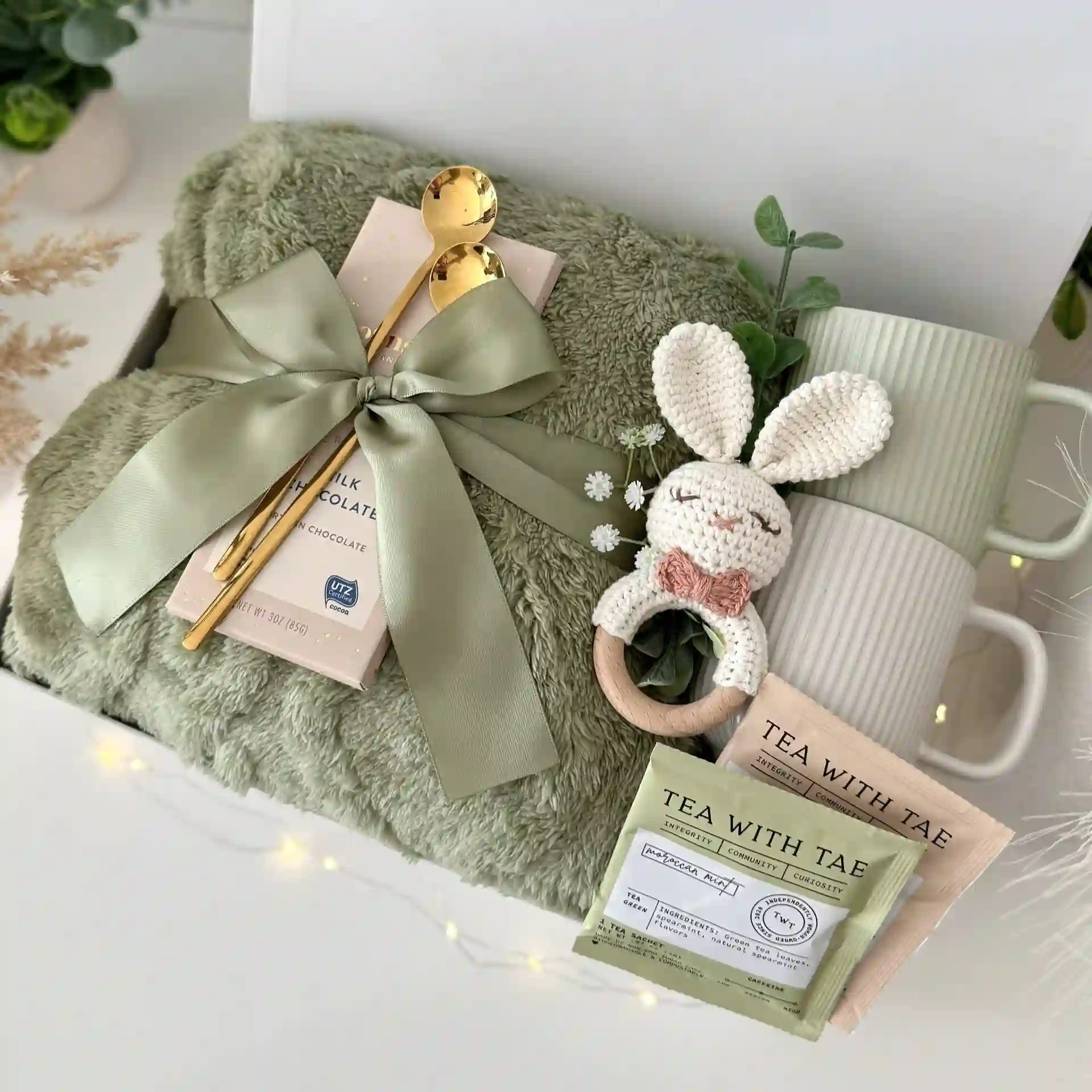 Gift box with two ribbed mugs, tea sachets, a milk chocolate bar and two gold spoons on a sage fleece blanket tied with a wide satin bow