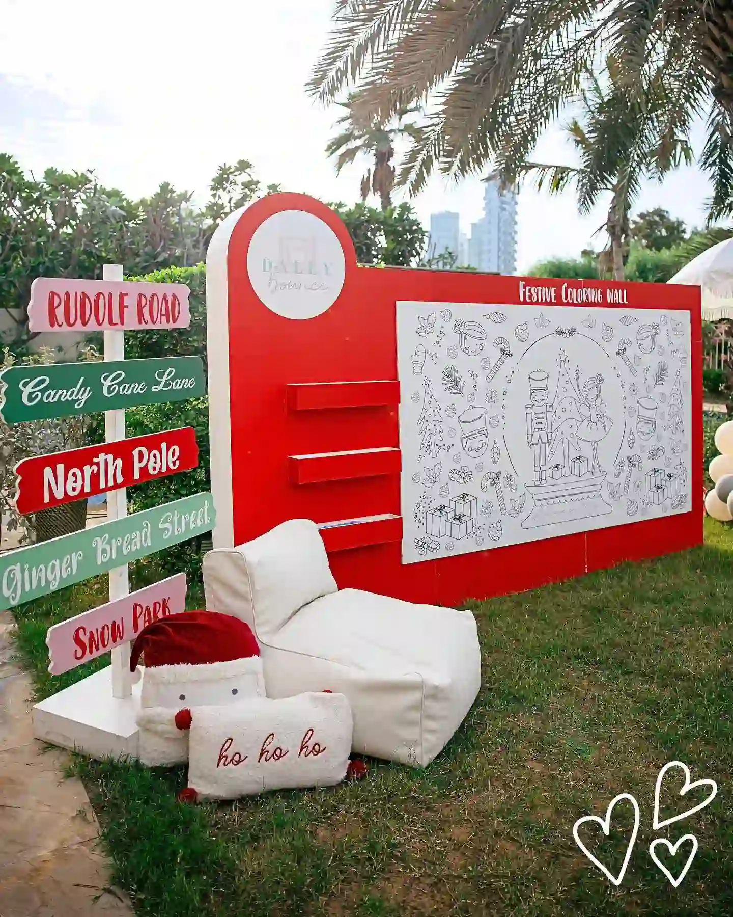 A red outdoor coloring wall with a large Christmas line drawing, marker shelves, a candy colored North Pole signpost, a white bean bag, and Santa pillows on the grass.