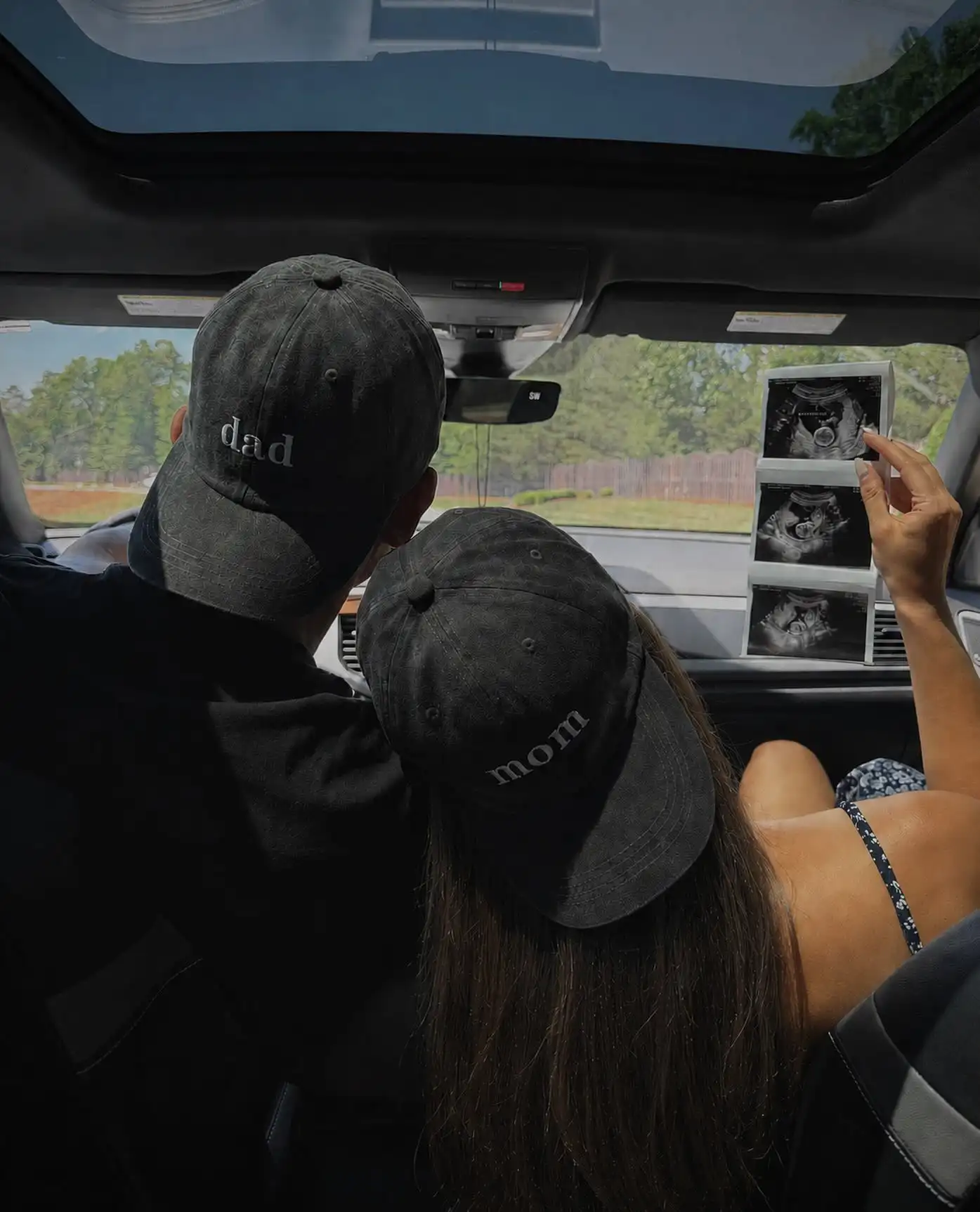 Couple sitting close in a parked car wearing dark washed caps reading dad and mom, holding a strip of three scan photos up against the windshield