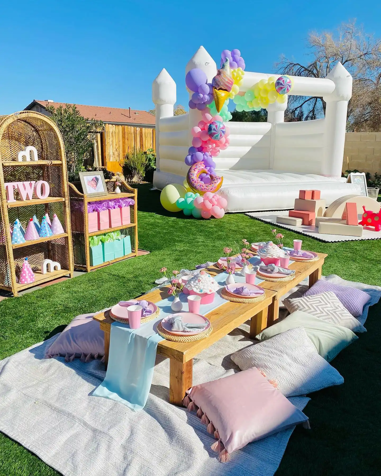Backyard birthday with a white bouncy castle and a pastel balloon column with ice cream, donut and candy foils, a soft play mat with foam blocks, a low table on cushions and party hats on rattan shelves beside a big TWO