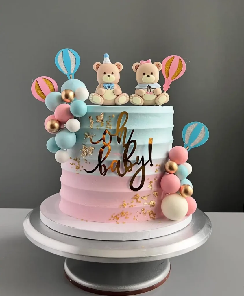 Tall ombre cake fading from blue to pink with two teddy bears, hot air balloon toppers, pastel and gold balls, and a gold oh baby sign