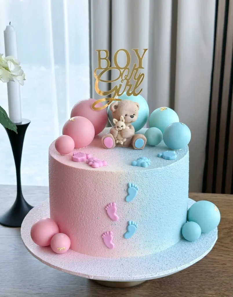 Round cake with a soft velvet finish blending pink to blue, with tiny footprints, pastel spheres, a bear holding a bunny, and a gold boy or girl topper