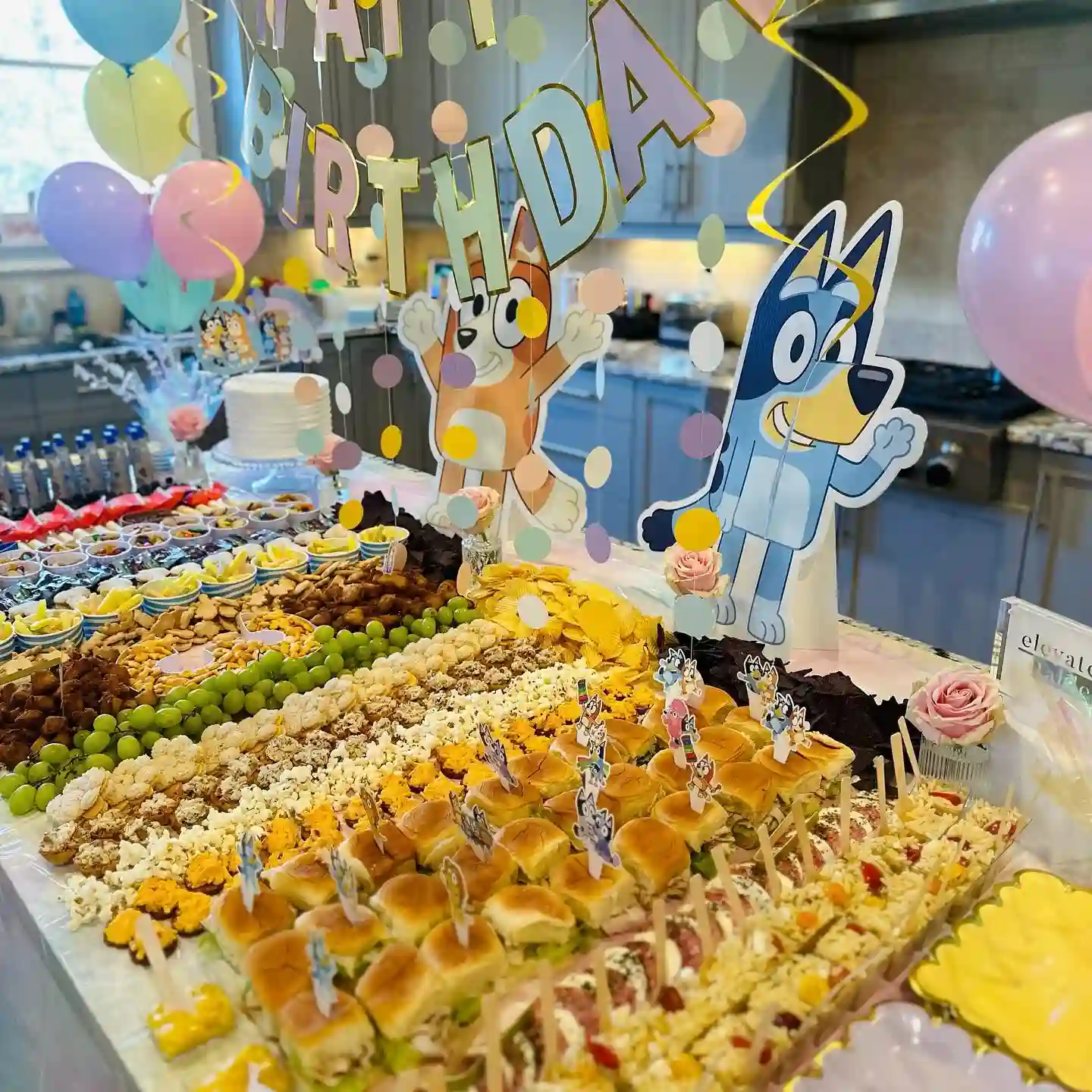 Pastel Bluey birthday table with stripes of grapes, popcorn, biscuits and chips and mini rolls with character picks