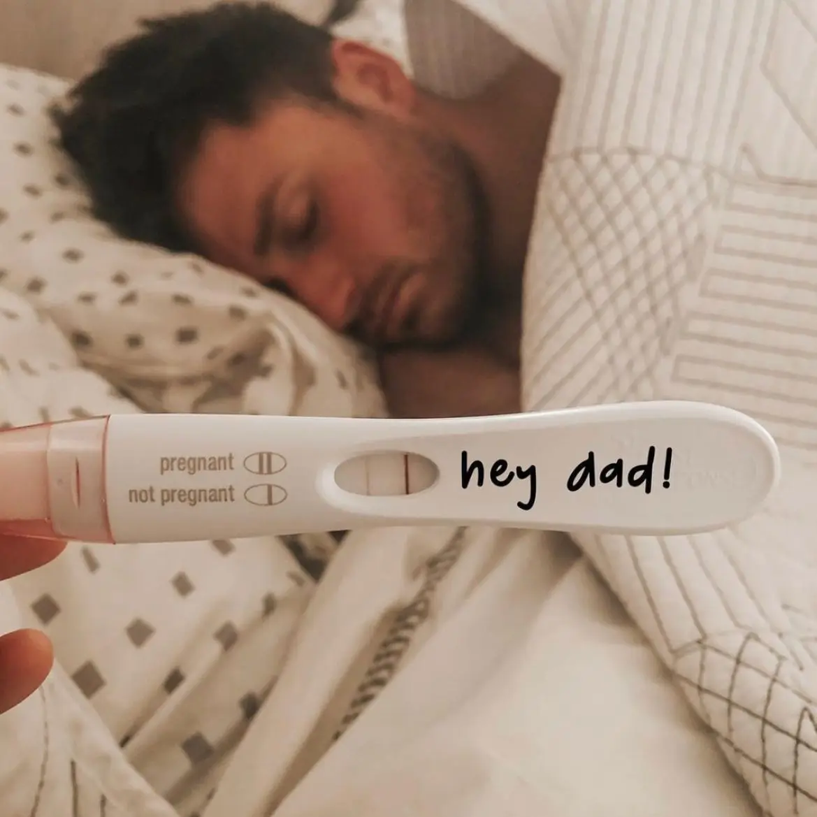 Pregnancy test with hey dad written in black marker on its handle, with a man asleep and blurred on a polka dot pillow behind it