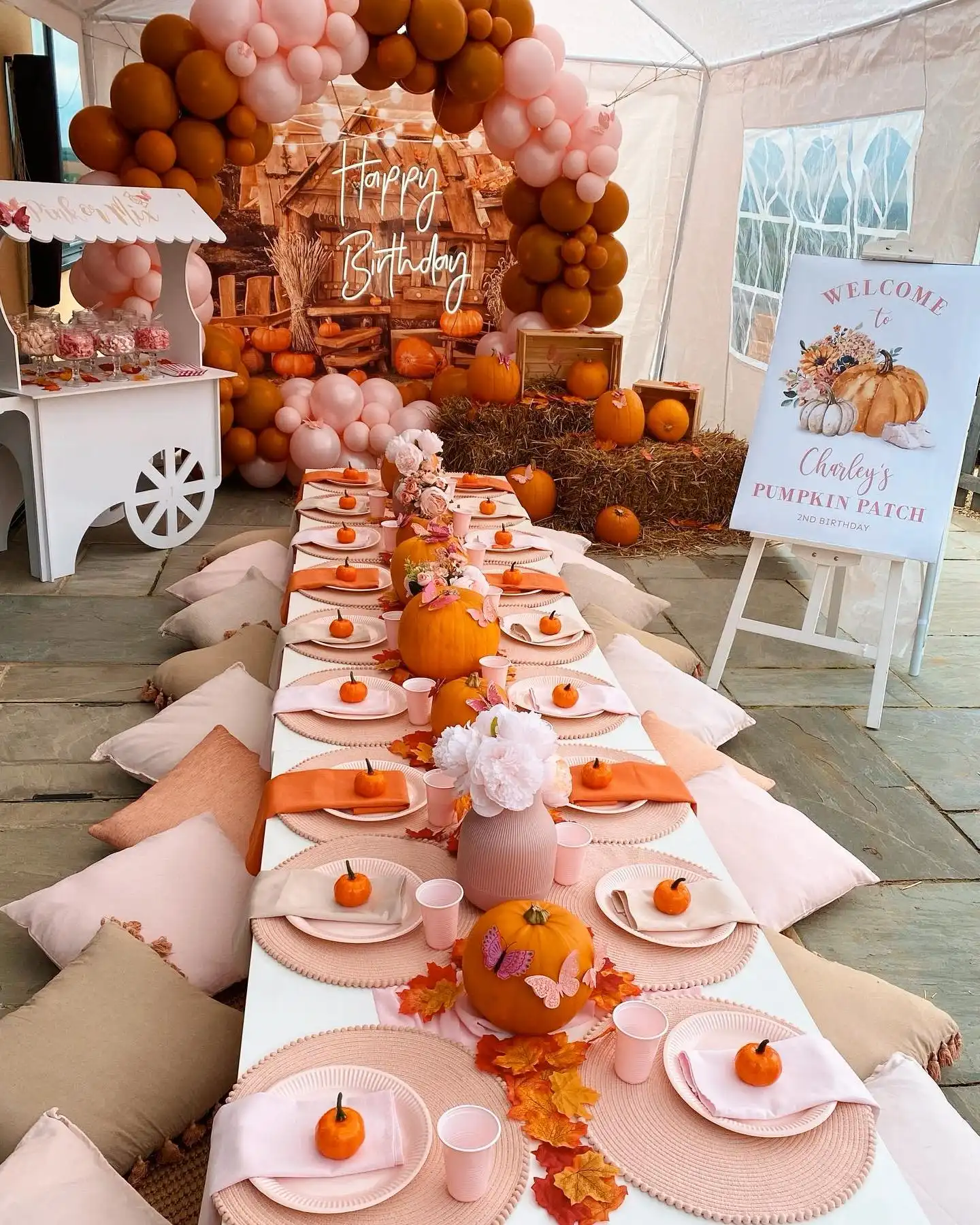 Pumpkin patch birthday with blush and burnt orange balloons around a rustic wooden cabin backdrop with a glowing Happy Birthday sign, hay bales, crates, a welcome sign on an easel, a white candy cart and a long table of pumpkins with floor cushions
