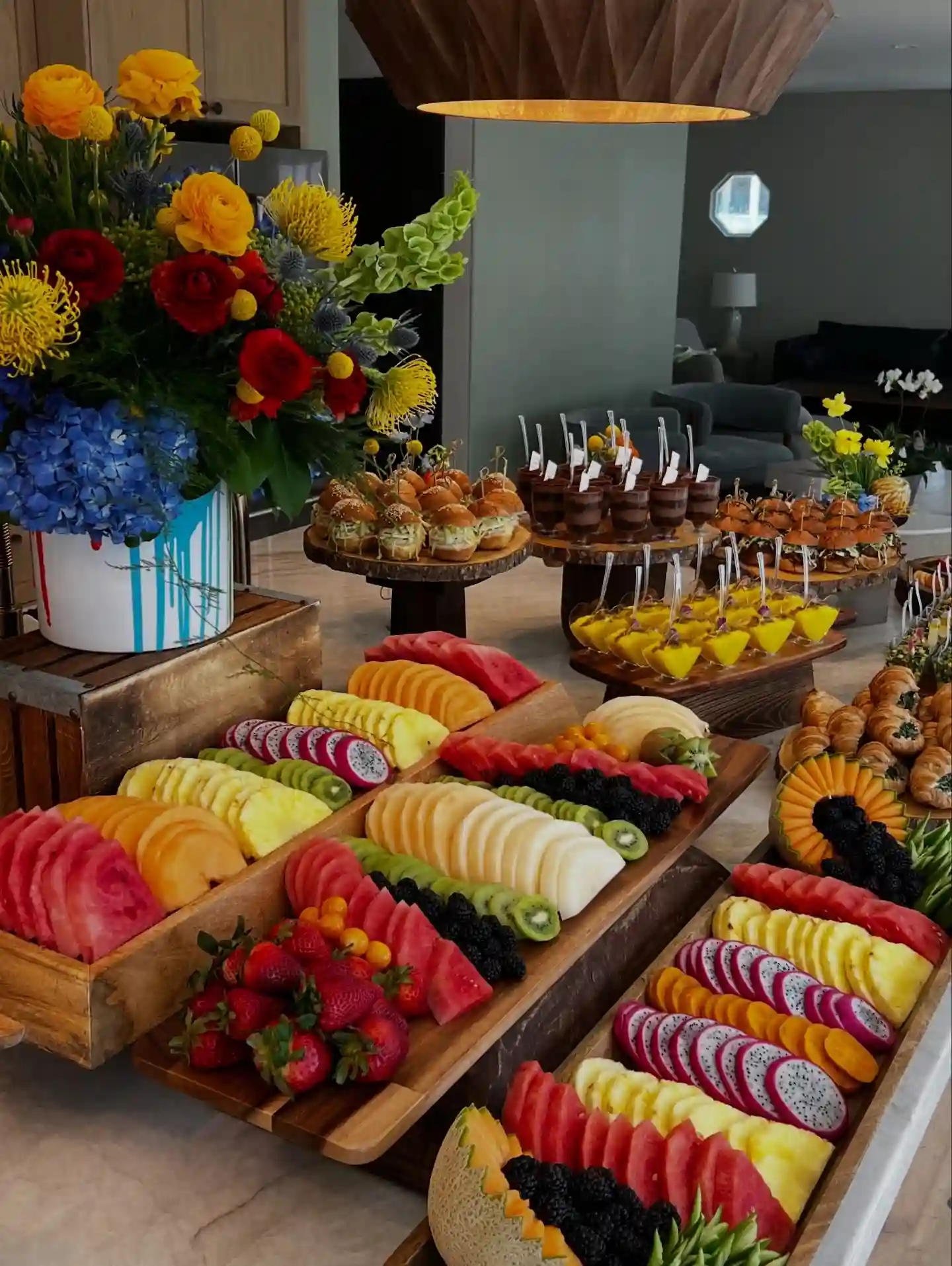 Wooden boards of sliced watermelon, pineapple, cantaloupe, dragon fruit and kiwi laid in colour rows beside mini sliders and dessert cups