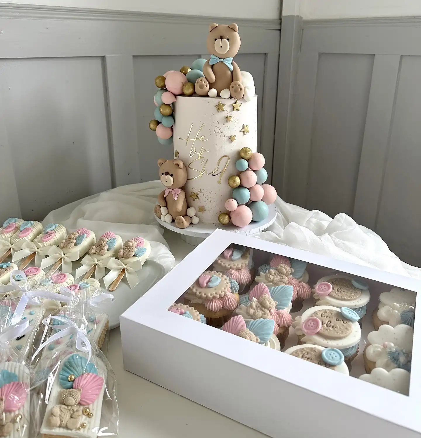 Tall blush gender reveal cake with two teddy bears, gold he or she sign and stars, beside treats on sticks and a box of decorated cupcakes