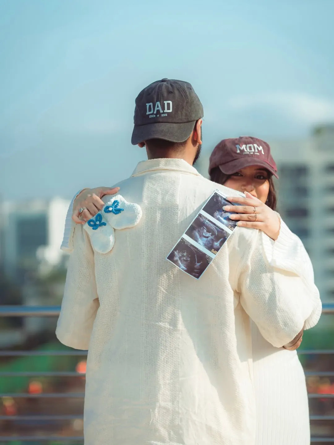 Man seen from behind in a DAD cap being hugged by a woman in a MOM cap, with ultrasound photos and white knit booties with blue bows against his cream jacket
