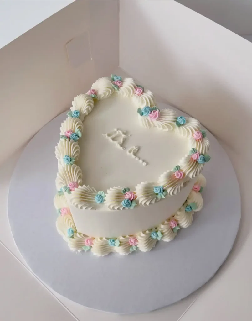 Cream heart shaped cake with shell piping, small pink and blue rosettes with mint leaves, and a short piped message that trails off in dots