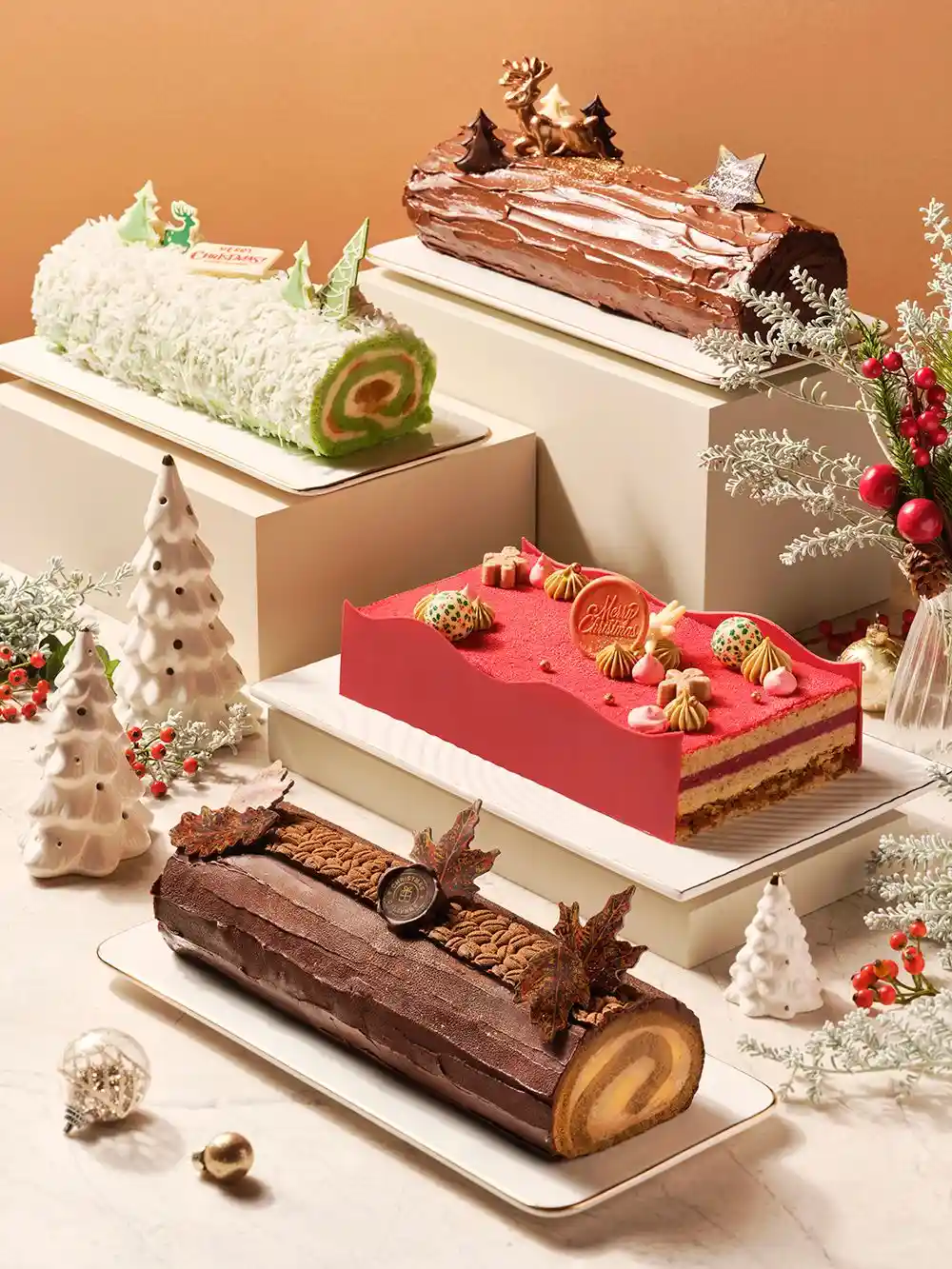 Four Christmas cakes on cream blocks of different heights: a snowy coconut roll with green trees, a dark chocolate log with a gold reindeer, a red layer cake with meringues and a chocolate log with leaves