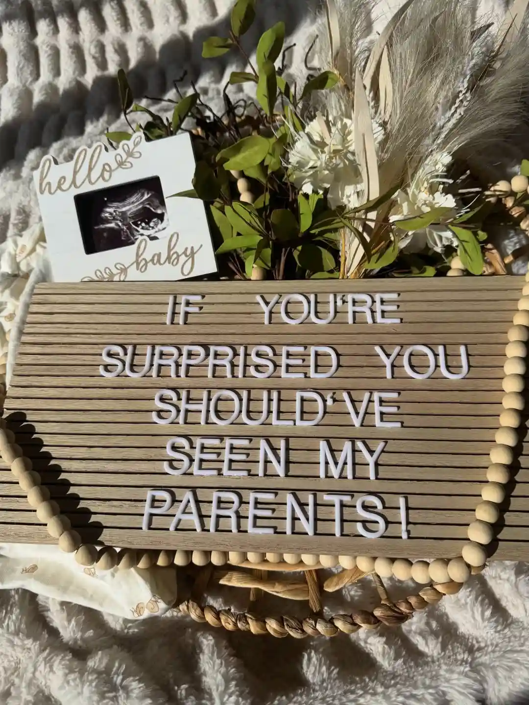 Wooden letter board reading "If you're surprised you should've seen my parents" beside a Hello Baby ultrasound frame, dried flowers and a wooden bead garland on a fluffy white blanket