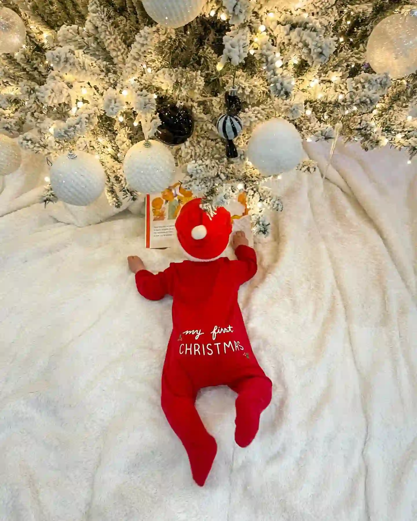 Baby on a fluffy white blanket in a red sleeper and Santa hat with a bright picture book, seen from above under a Christmas tree hung with white and black ornaments