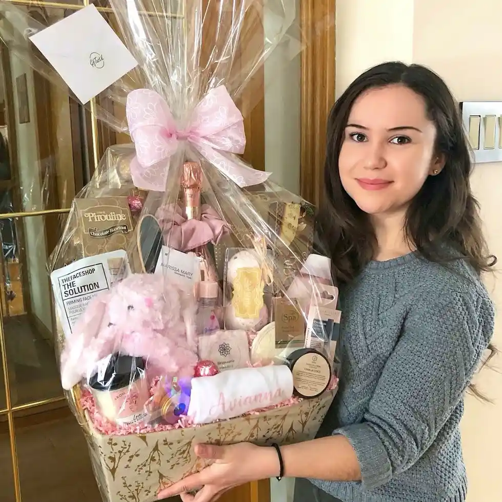 Spa gift basket wrapped in cellophane with a pink bow, holding a sheet mask, a chocolate hazelnut box, spa bath items, a pink scrunchie, a vanilla tin, a pink plush bunny and a rolled blanket with a stitched name