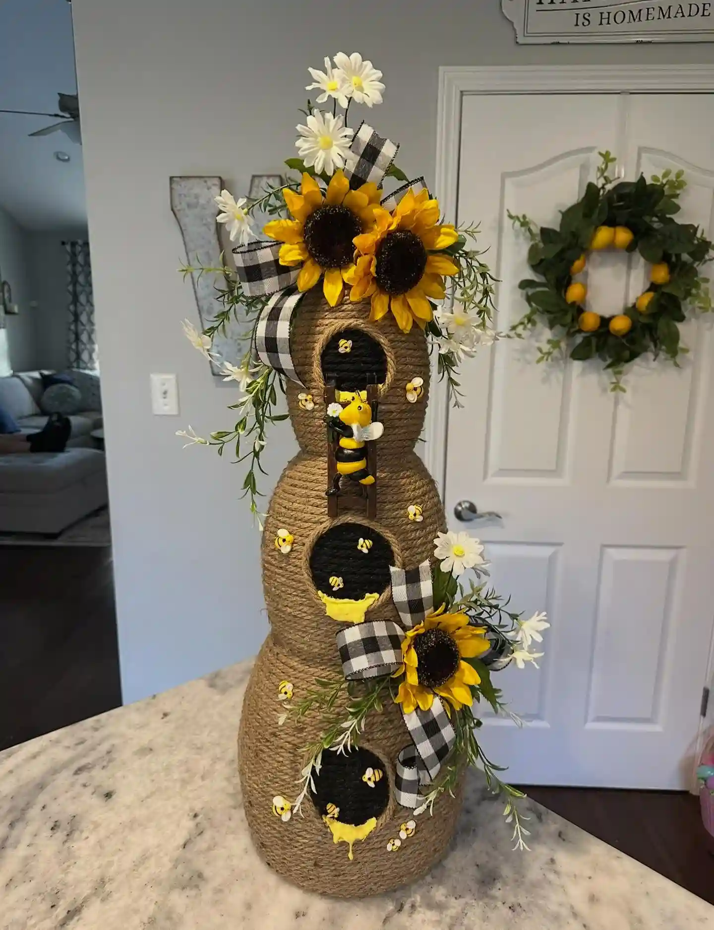 Three stacked round forms wrapped in jute twine as a beehive with black round doors, tiny bees, yellow honey drips, a small ladder and sunflowers, white daisies, greenery and black and white check ribbon