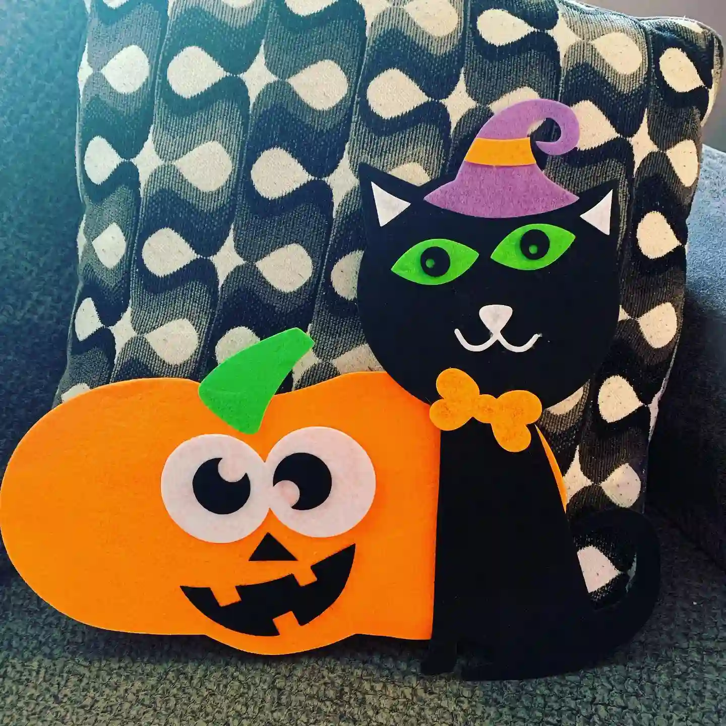 Felt craft of a grinning orange pumpkin with big white eyes and a black cat in a purple witch hat with green eyes and an orange bow tie sitting together