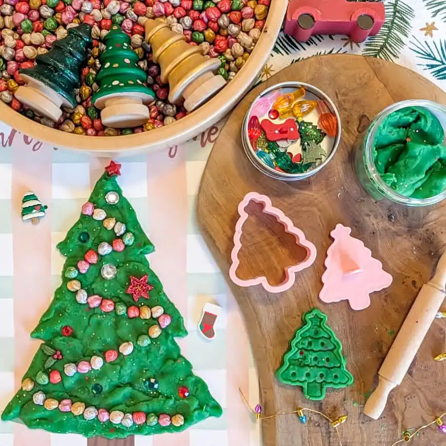 A green playdough Christmas tree decorated with colorful beads and a red star, beside tree shaped cutters, a rolling pin, a jar of green dough, and a bowl of beads with small wooden trees.
