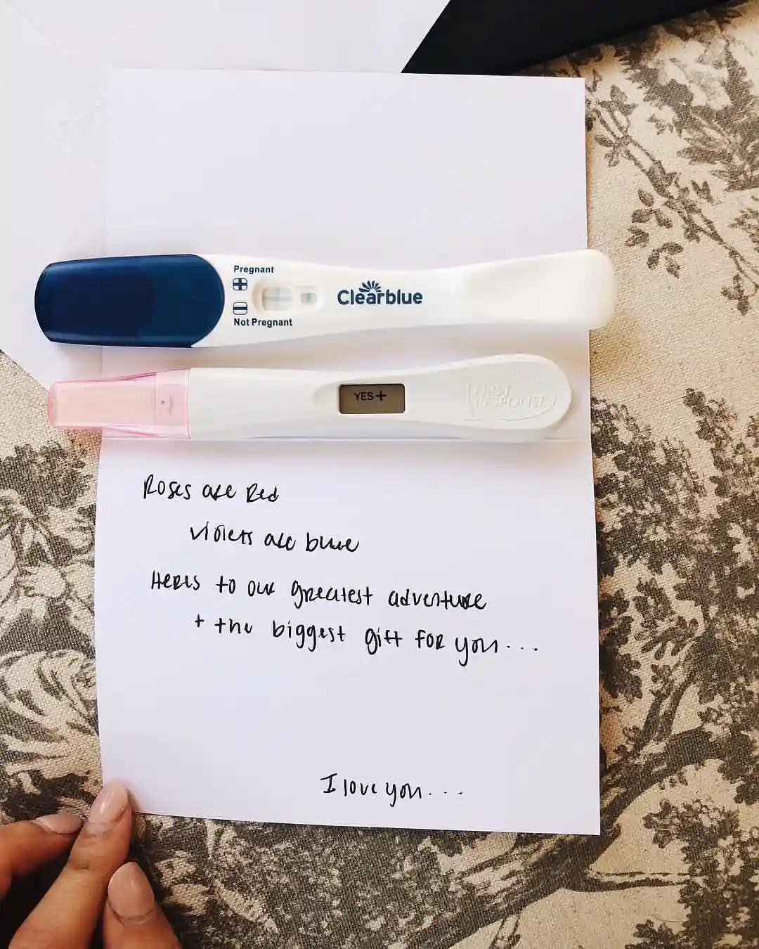 Open white card on patterned fabric with two pregnancy tests across the top and a handwritten rhyme below ending with I love you