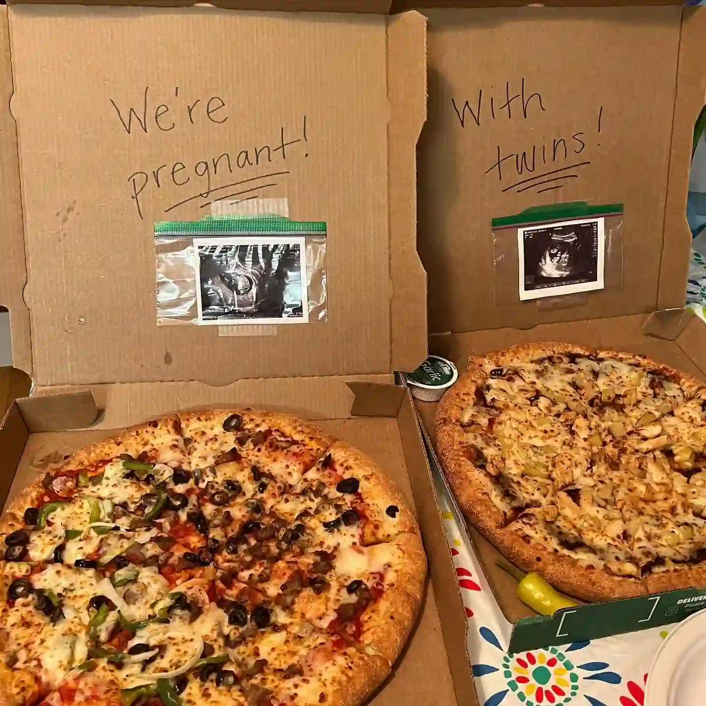 Two open pizza boxes with handwritten lids reading "We're pregnant!" and "With twins!", each with an ultrasound photo taped inside a zip bag, above a veggie pizza and a chicken pizza
