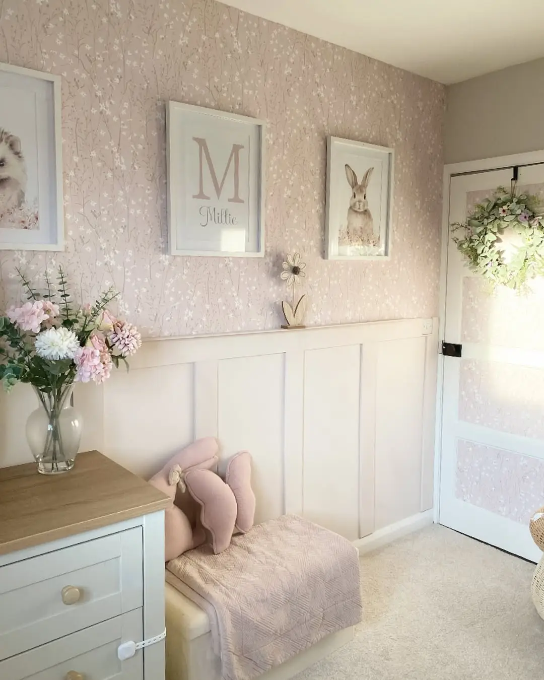 Blush floral wallpaper above white wall panelling in a baby girl's nursery with framed animal prints