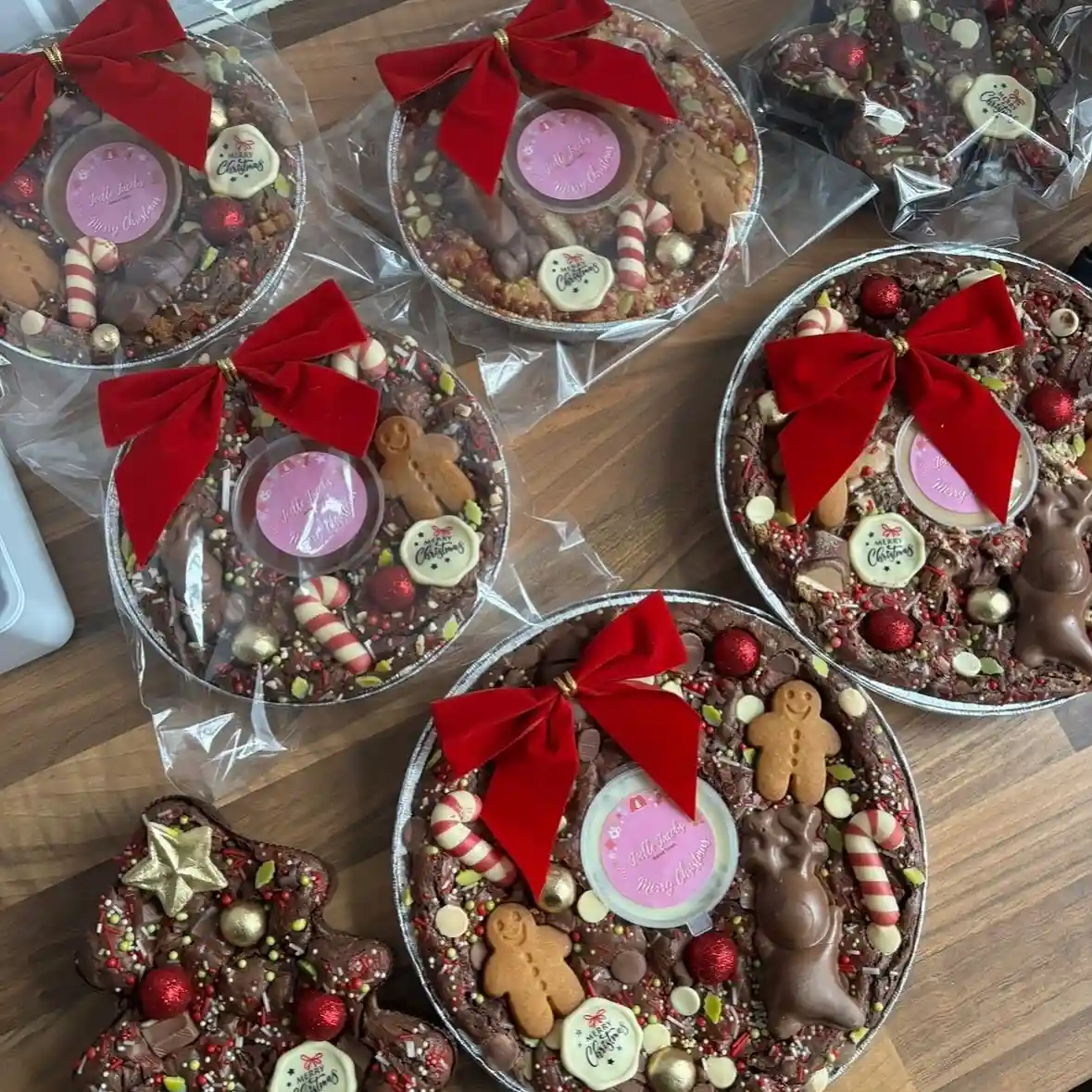 Round brownie trays covered in chocolate pieces and sprinkles, topped with candy canes, gingerbread men, baubles and a Merry Christmas plaque, with a big red velvet bow