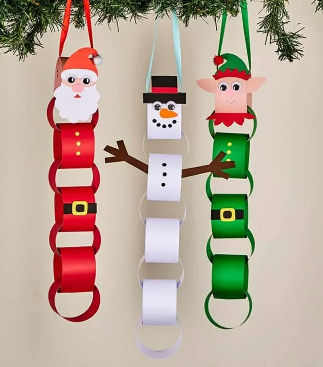 Three hanging paper chains shaped like Santa, a snowman, and an elf, each with a paper face on top, hung from ribbon loops under a pine branch.