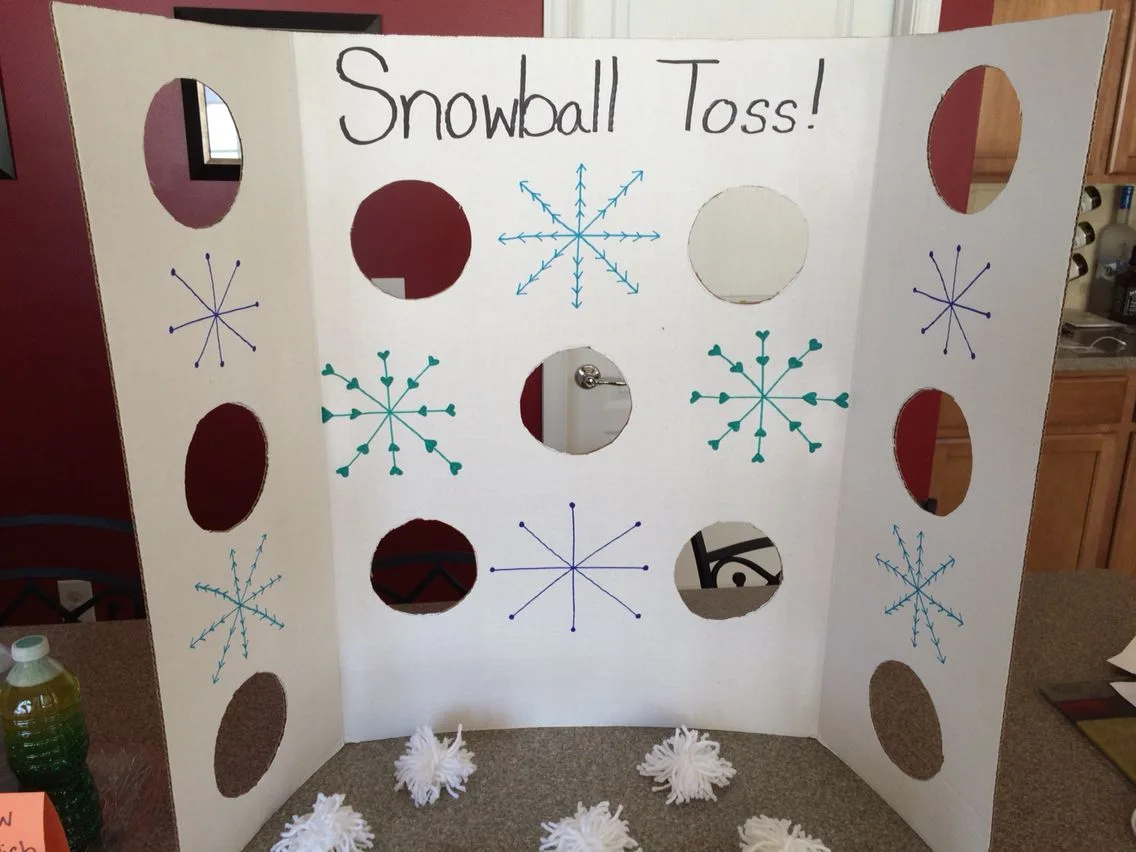 A white tri-fold board with round holes, hand drawn snowflakes, and the words Snowball Toss, with white yarn pom pom snowballs on the counter below.