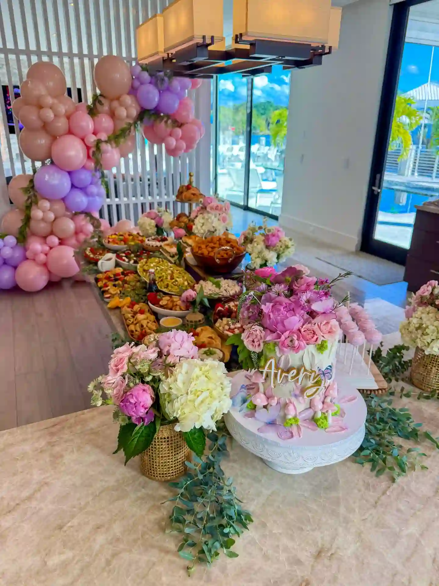 Garden-style birthday table with pink and purple balloon garland, flower arrangements, a floral cake and bowls of mixed party food