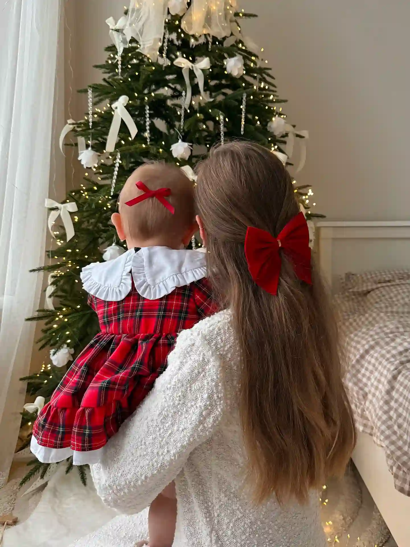 Mom holding baby in a red and green tartan dress with a white ruffled collar up to a Christmas tree hung with white ribbons, flowers and red bows